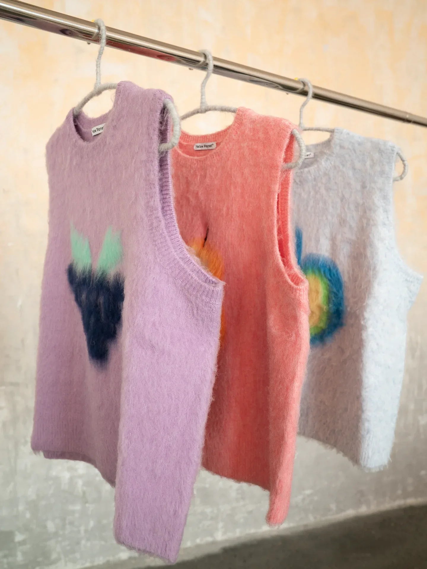 A close-up side view of the purple, pink pear, and light blue knit vests from the Yellow Market Series hanging on a rack.