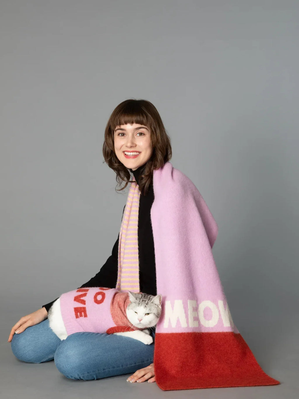 A woman modeling the CAT-Sophy "MEOW" theme oversized scarf in soft pink, brick red, and yellow-pink stripes while holding a cat.