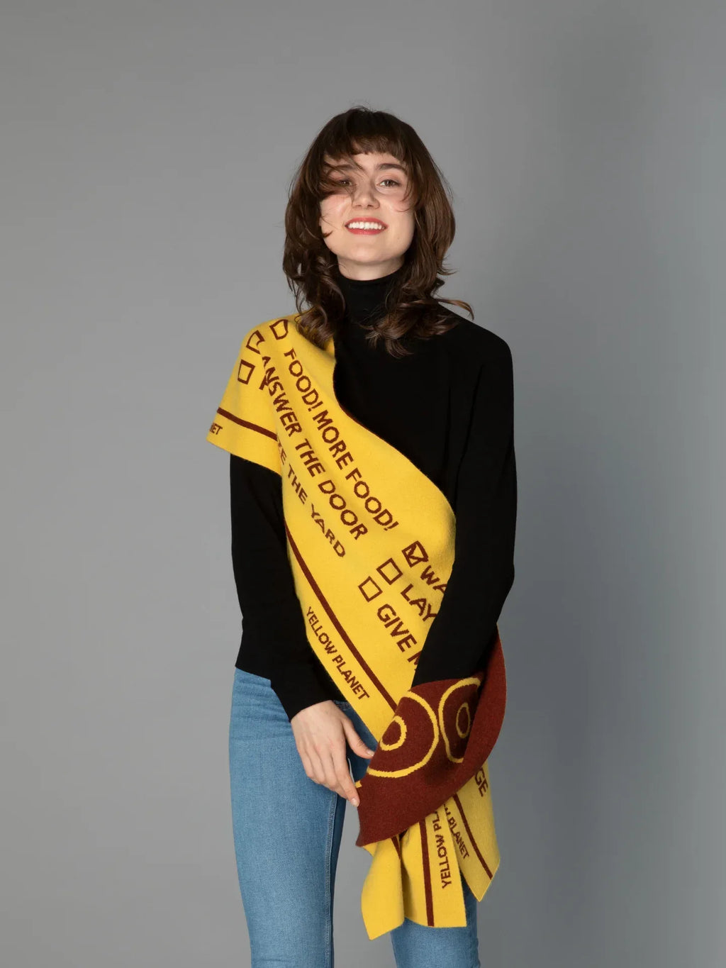 A woman modeling the DOG-Sophy "WOOF" theme reversible scarf in warm yellow and brown, draped over a black turtleneck.