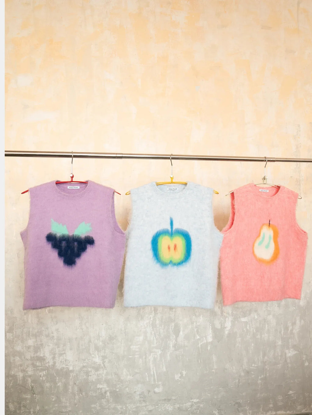 Multiple fruits brushed knit vests hanging in a row, including the Pink Pear vest next to purple and blue options. part of the Yellow Market collection.