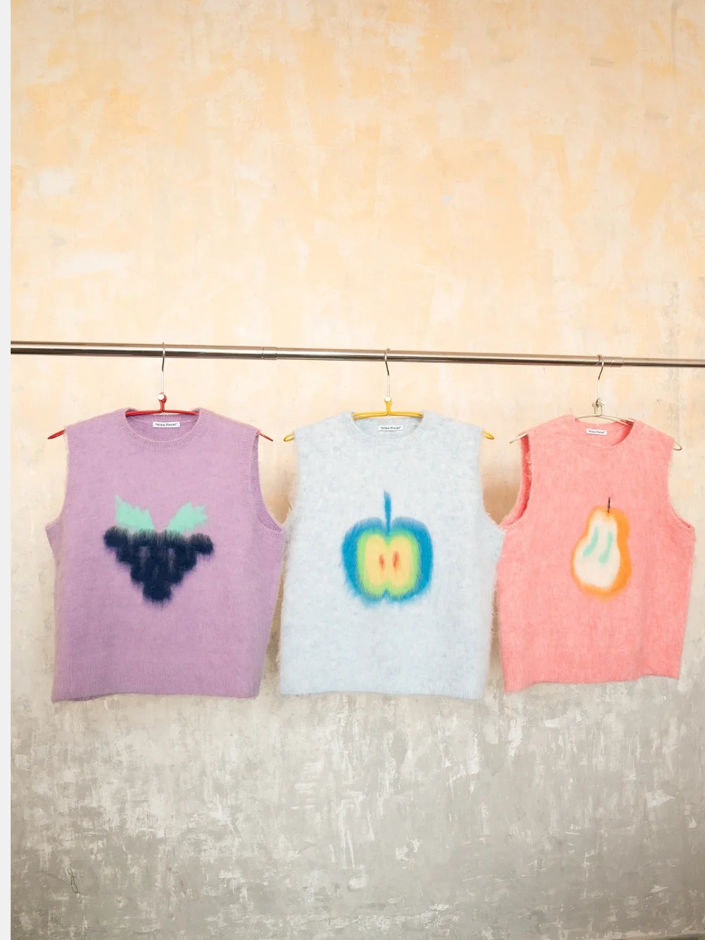 Multiple fruits brushed knit vests hanging in a row, including the Pink Pear vest next to purple and blue options. part of the Yellow Market collection.