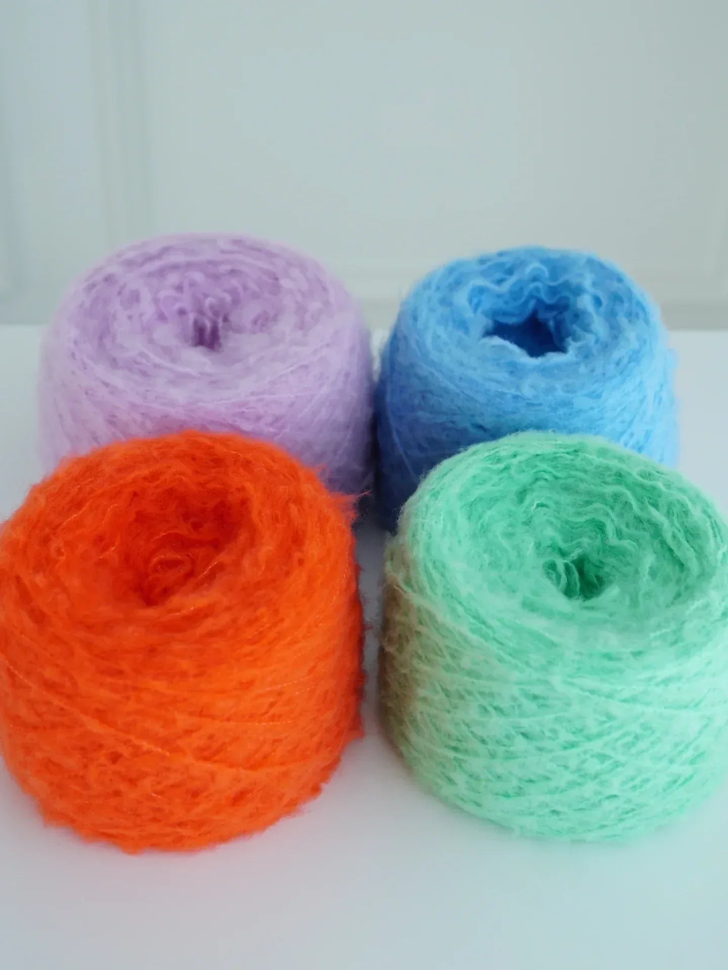 Four vibrant rolls of fuzzy wool yarn in purple, blue, orange, and mint green used for the Mochi BonBon collection.
