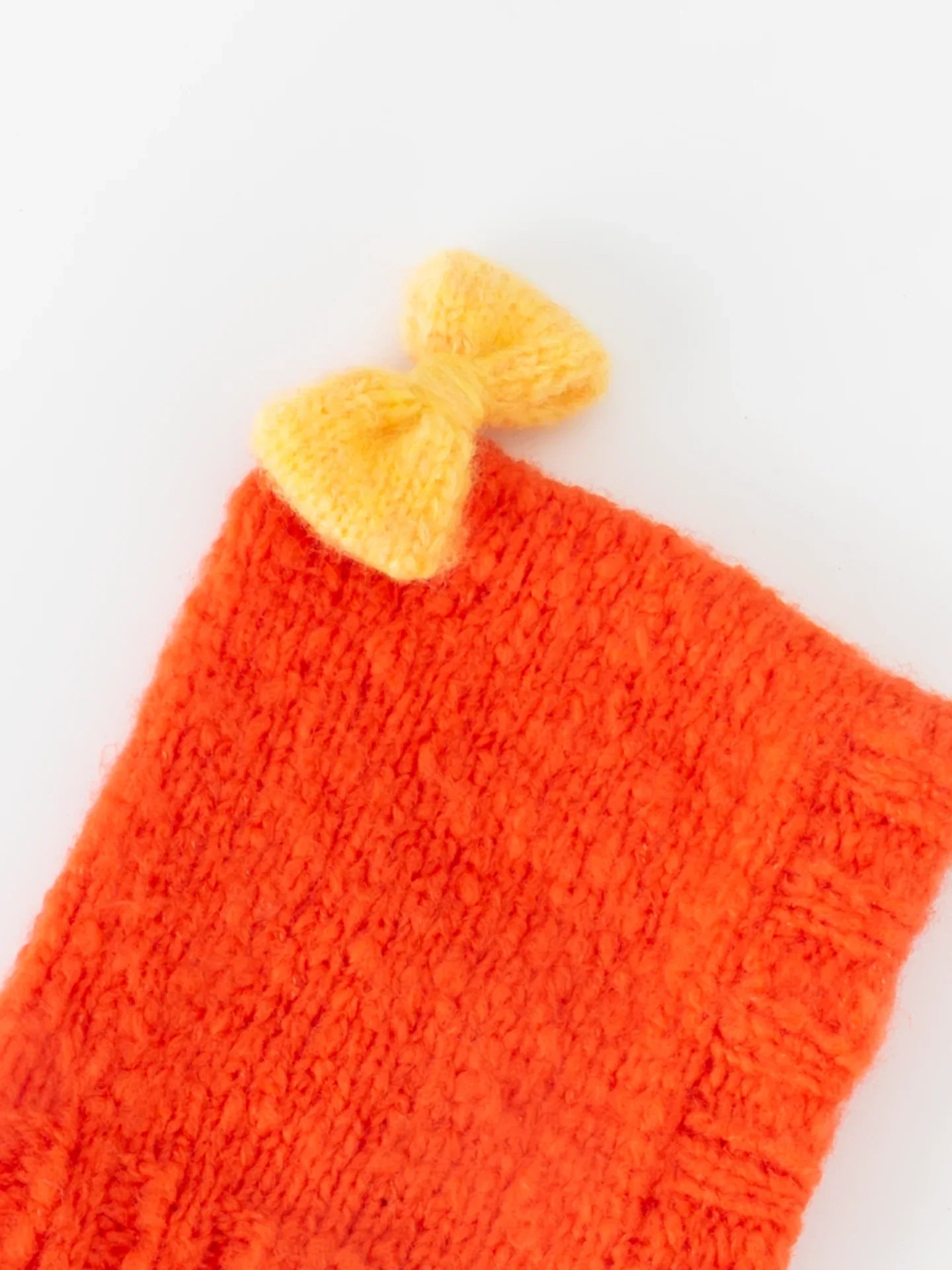 Close-up shot of the hand-knitted warm yellow bow accent on the crown of the persimmon red wool beanie.