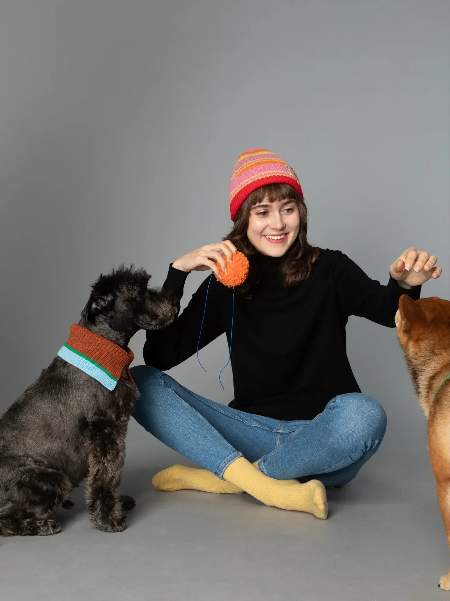 A woman wearing the Pink Stripes beanie while sitting with a black dog wearing a matching striped pet collar/snood.