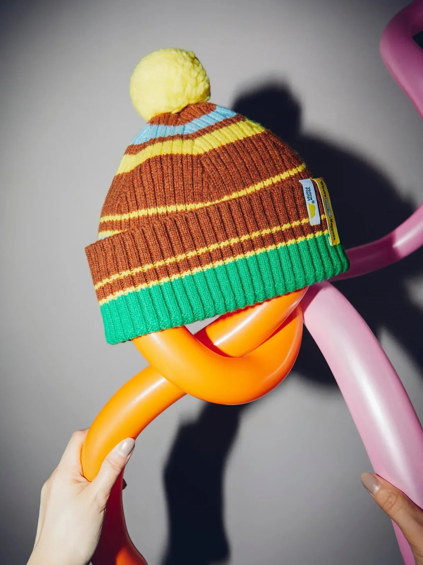 Close-up of the striped beanie styled with its bright yellow detachable pom-pom held against a playful balloon background.
